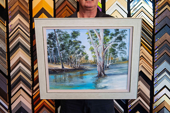 Picture Framer Scott holding a custom framed original landscape painting in the Sunset Framing showroom in Port Macquarie.