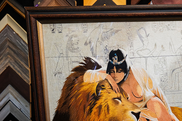 Close-up of custom framed Egyptian-inspired textile art showing detailed fibre work of a figure resting against a lion, surrounded by hieroglyphic imagery in a dark timber-look frame with gold inner d