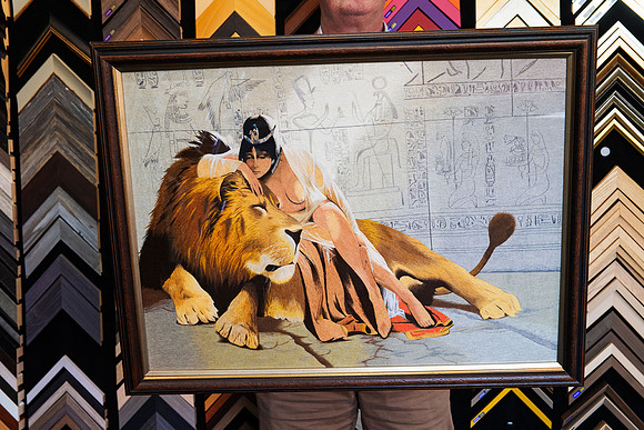 Detailed Egyptian-inspired textile artwork featuring a reclining lion and figure with hieroglyphic imagery, custom framed in a dark brown timber-look picture frame.