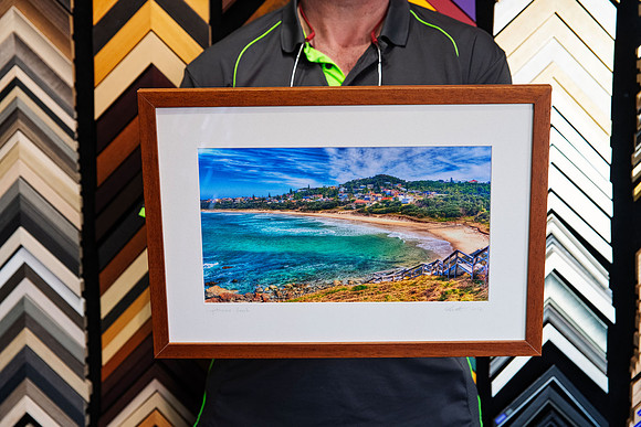 Framed colour photograph of Lighthouse Beach Port Macquarie featuring turquoise ocean, sandy beach and coastal headland, presented with a wide white mat and warm timber custom picture frame.