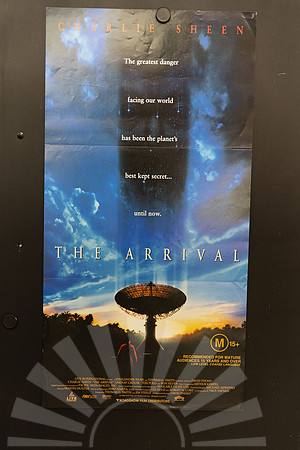 The Arrival Australian Daybill with a shadowy alien face amid blue clouds and stars, above a radio telescope at sunset and a small red-lit figure.